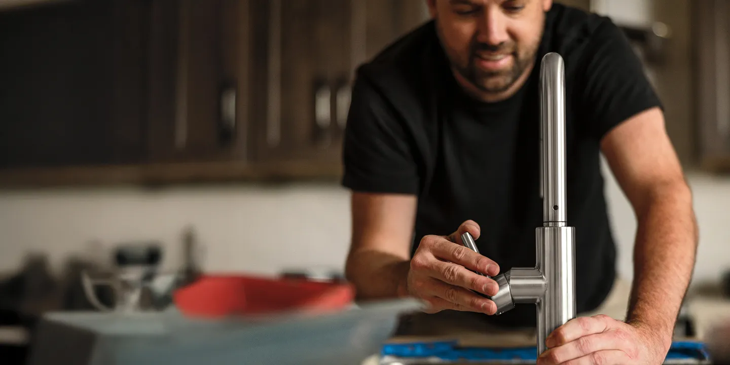 Plumber/installer in kitchen with dark cabinets (background) installing chrome single-handle Delta faucet (foreground).